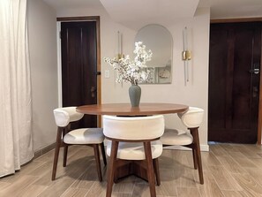 A dining area with a round wooden table, four white upholstered chairs, a vase with white flowers, a mirror, and two doors. - Studio Near Wrigley • Sleeps 4 • Games + Fireplace (Chicago)