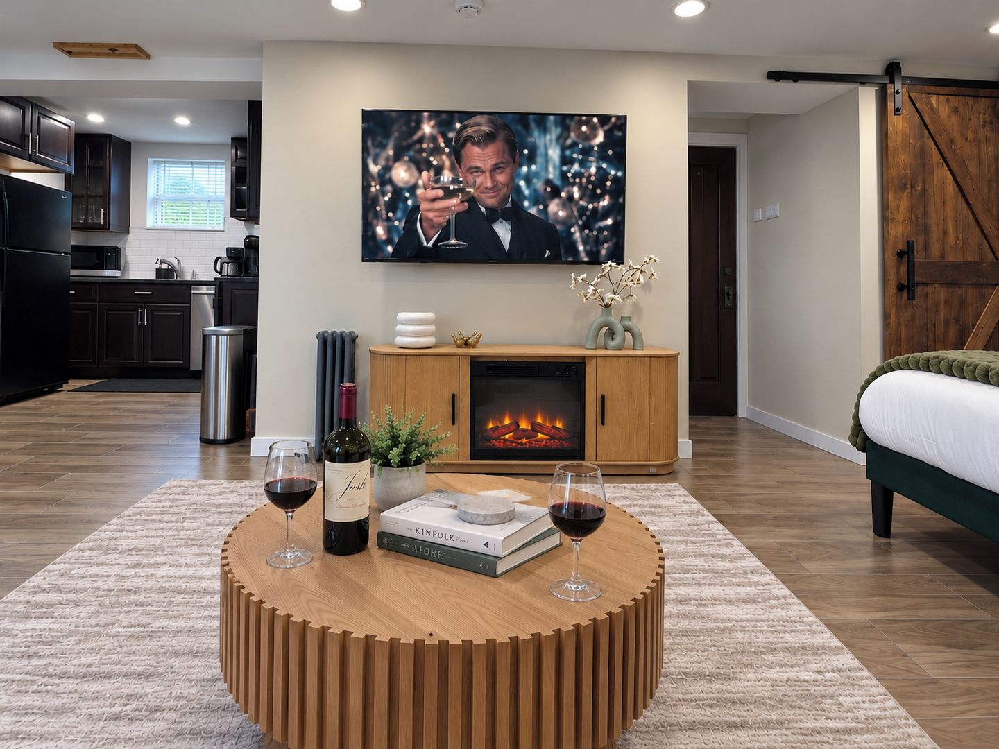 Un salon moderne avec une cheminée, une table basse ronde sur laquelle sont posés des livres et des verres à vin, un téléviseur au-dessus de la cheminée et une cuisine en arrière-plan.