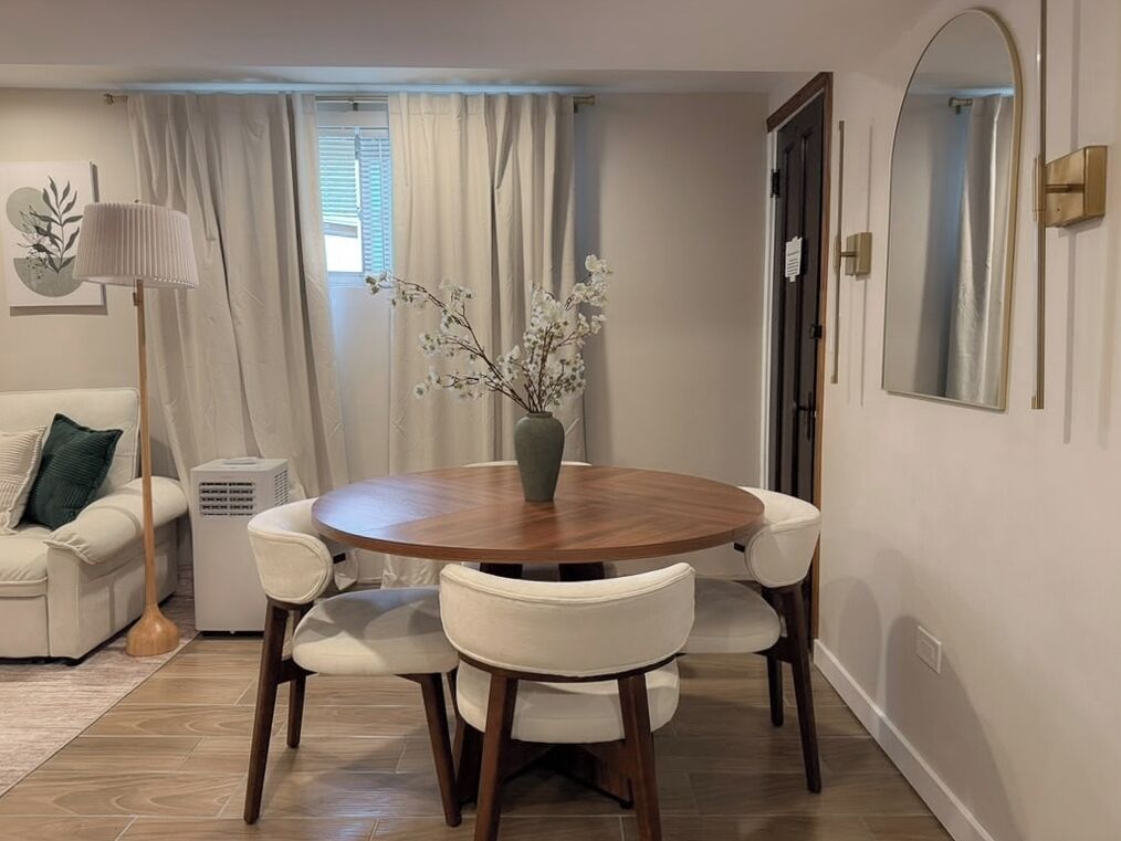 Un coin repas avec une table ronde en bois, des chaises à revêtement blanc, un vase avec des branches, une lampe sur pied et un purificateur d’air blanc.
