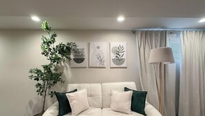 Un salón con sofá blanco, cojines verdes, una planta en maceta y cuadros en la pared.