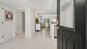 Un couloir mène à un espace de vie comprenant un canapé et une table basse, une porte ornée d’un panneau décoratif et une plante en pot.
