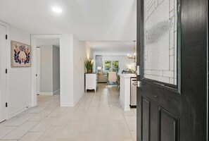 Un pasillo que conduce a una sala de estar con sofá y una mesa de centro, una puerta con panel decorativo y una planta en maceta.