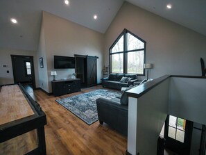A spacious living room with a large window, a flat-screen TV, a dark wooden entertainment unit, and a cozy sofa. - Relax at Tees, Trees & Skis: a 4-bedroom retreat with hot tub and golf views on Mountaintop. (McHenry)