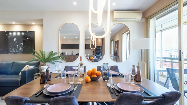 Un coin repas avec une table en bois dressée pour quatre personnes, entourée de chaises, avec une cuisine en arrière-plan et une grande fenêtre.