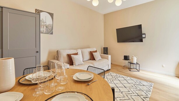 A dining area with a round table set for four, a sofa in the background, a television mounted on the wall, and a door with a view of the Eiffel Tower. - Bright Cosy Suite Paris 16th Steps From Seine (Paris)