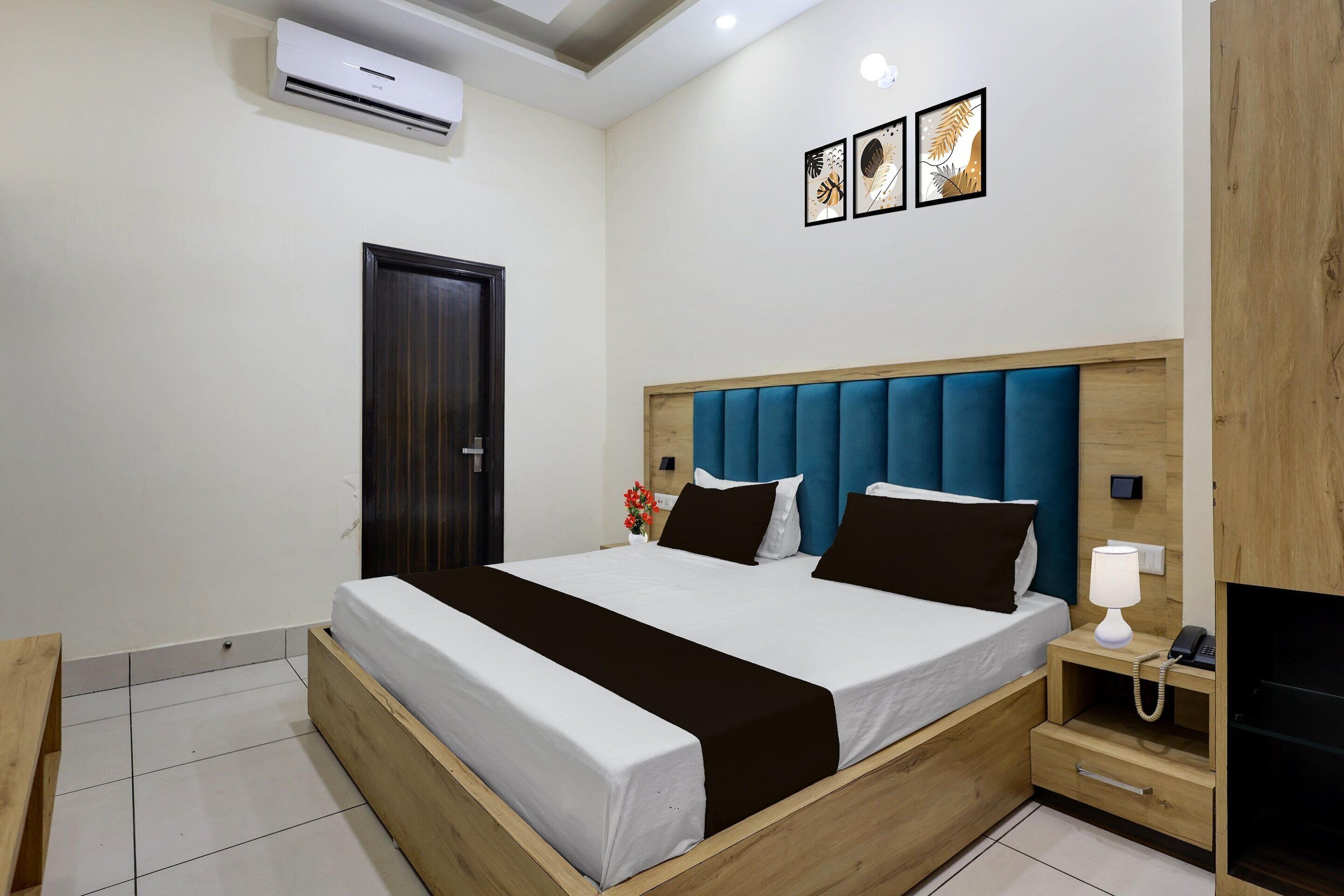 A modern hotel room with a wooden bed frame, a blue upholstered headboard, a bedside table with a lamp, and a wall-mounted air conditioner.