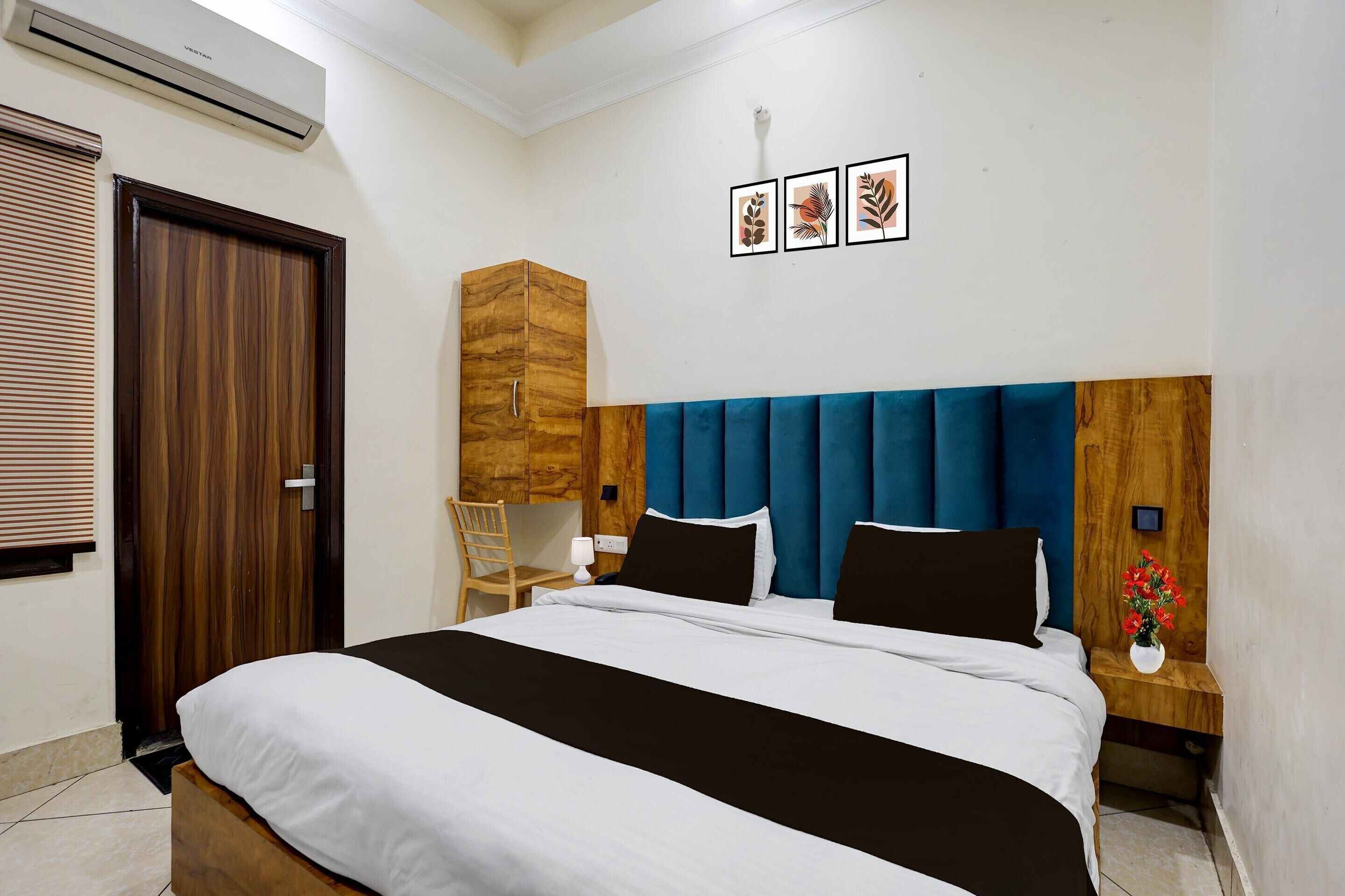 Een slaapkamer met een bed, een houten garderobe, een stoel, een nachtkastje met een vaas, en ingelijste foto’s aan de muur.