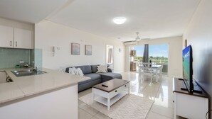 A modern living room with a sofa, coffee table, and TV. There is a kitchen area with a sink and a dining area with a table and chairs. - Platinum Apartments -  Unit 22 (Bargara)