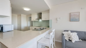 A modern kitchen with a central island, white cabinetry, and a grey sofa in the living area. - Platinum Apartments -  Unit 22 (Bargara)