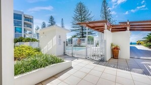 A gated pool area with a white fence, a small building with a sign, and a view of a yellow car on a road. - Dual Living, Pool, Gym, Sleeps 8, Parking (Bargara)