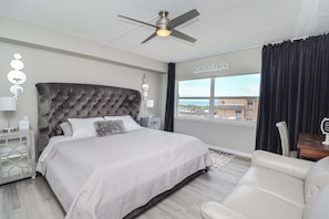 Ein Schlafzimmer mit einer gesteppten Kopfteil, einem Bett mit einem gemusterten Bettüberwurf, einem Deckenventilator, einem Nachttisch mit Lampe, einem Beistelltisch mit Lampe und Blick auf das Meer durch ein Fenster.