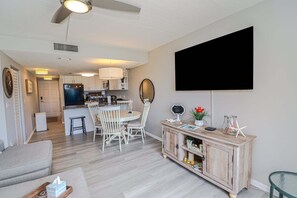 Ein modernes Wohnzimmer mit Flachbildfernseher, einem Essbereich mit rundem Tisch und Stühlen, einer Küchenzeile und einer kleinen Kommode mit Dekoration.
