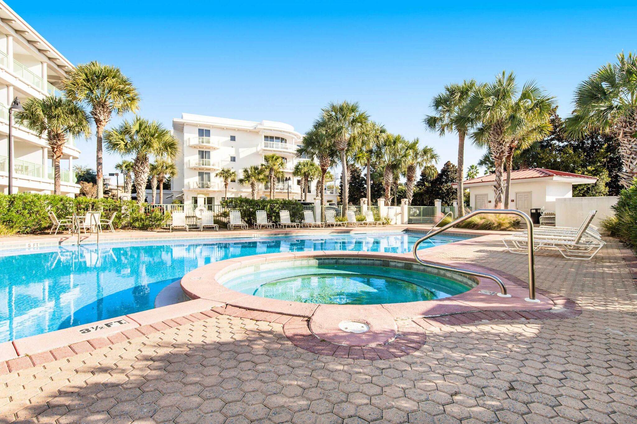 A swimming pool with a hot tub, surrounded by palm trees and lounge chairs, with a building in the background.