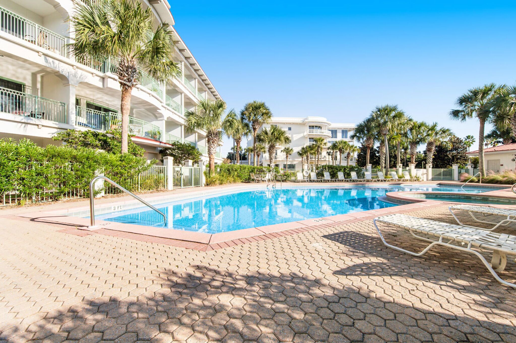 Ein Hotelpoolbereich mit klarem, blauem Pool, Liegestühlen, Palmen und einem mehrstöckigen Gebäude mit Balkonen.