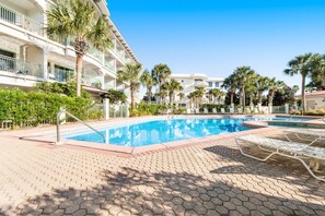 Un espace piscine d’hôtel avec une piscine à l’eau d’un bleu limpide, des chaises longues, des palmiers et un immeuble à plusieurs étages doté de balcons.