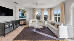 A living room with a fireplace, white sofas, a TV, and a large window with curtains. - GULF-FRONT 3BR Condo! Steps to Beach & Community Pool! (Seacrest)