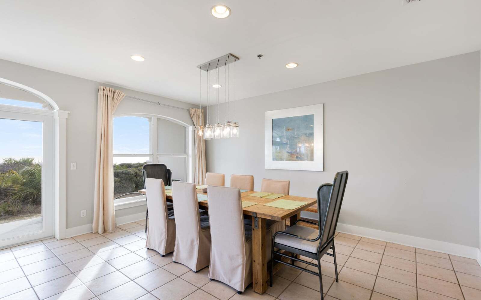 A dining area with a wooden table, beige chairs, a chandelier, and a large window with curtains.