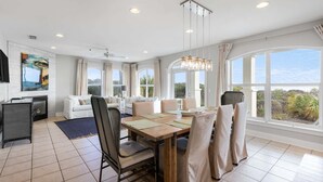 A spacious living area with a dining table, sofas, and large windows offering a view of the outdoors. - GULF-FRONT 3BR Condo! Steps to Beach & Community Pool! (Seacrest)