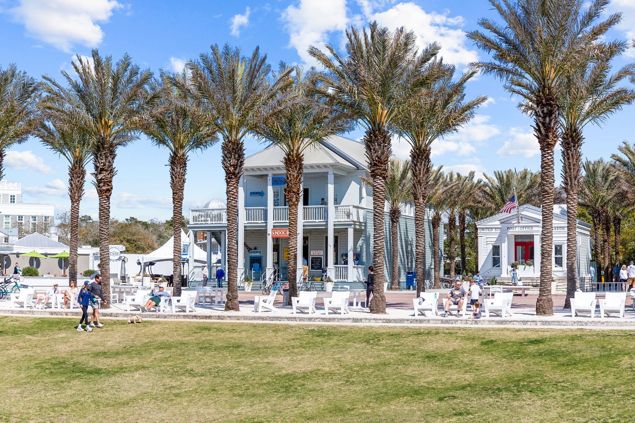 Ein Strandhotel mit weißer Fassade, Palmen und ein Strandbereich mit weißen Stühlen sowie ein kleines Gebäude mit der Aufschrift 'United States Post Office'.