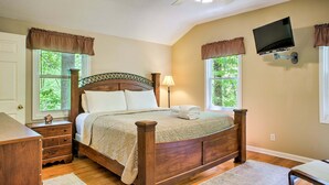 A bedroom with a wooden bed, a ceiling fan, a TV mounted on the wall, and two windows with curtains.