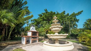 Um jardim com uma fonte central, espaço para churrasco e vegetação tropical exuberante.