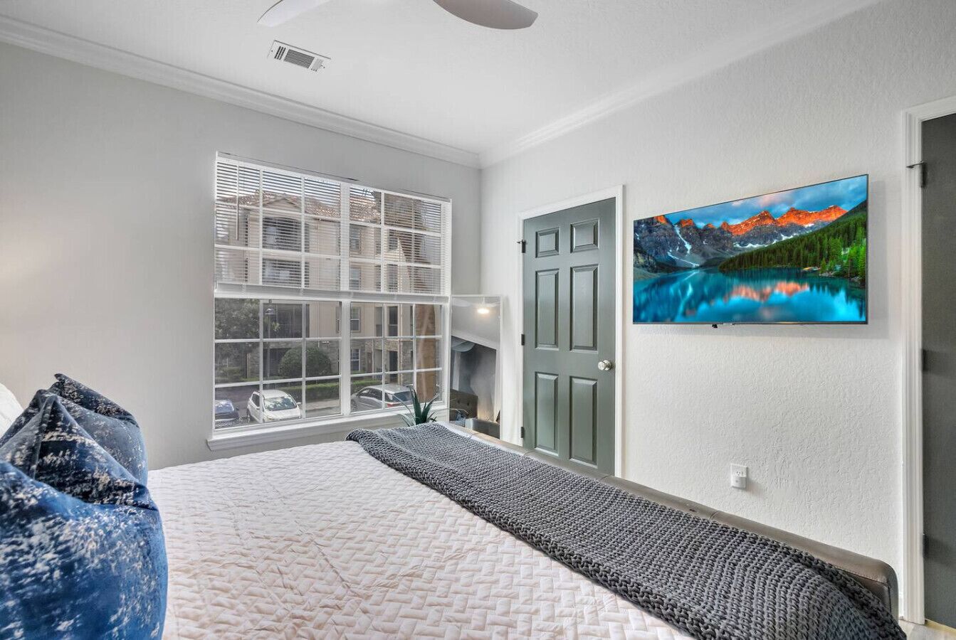 A bedroom with a bed, a window with blinds, a door, and a wall-mounted TV.