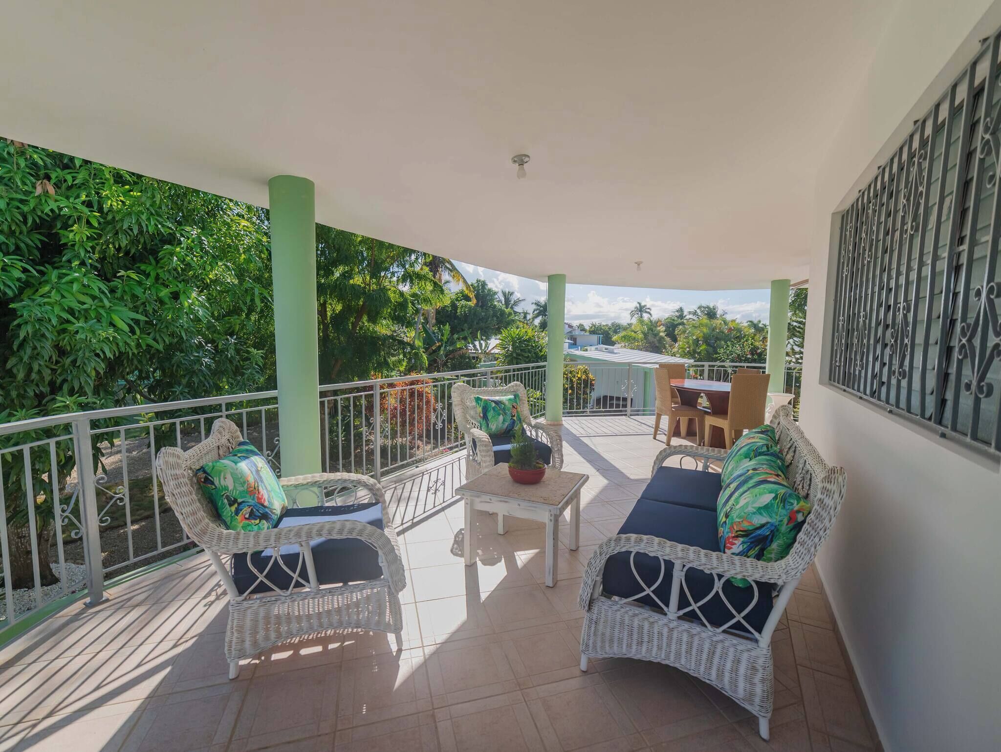 A covered balcony with wicker furniture, a small table, and a view of greenery and buildings.
