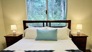 Ein ordentlich bezogenes Bett mit blauem Kopfkissen, zwei Nachttischlampen und einem Fenster mit Blick auf den Wald.