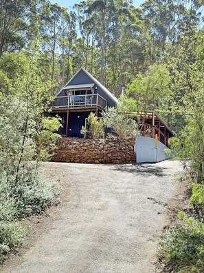 A two-story cabin with a balcony, surrounded by trees and a stone wall.