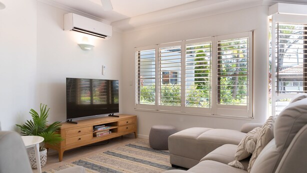 Una sala de estar con una televisión de pantalla plana, un mueble de madera, un reposapiés, un sofá color beige y un ventanal con persianas.