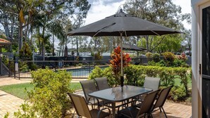 Un patio exterior con mesa y sillas, un paraguas grande y vistas a una piscina y árboles.