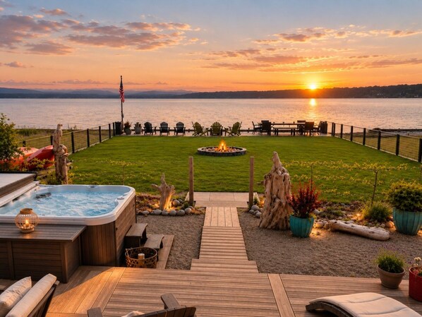 Una terraza con jacuzzi, asientos de madera y un pozo de fuego, con vista a un cuerpo de agua al atardecer.