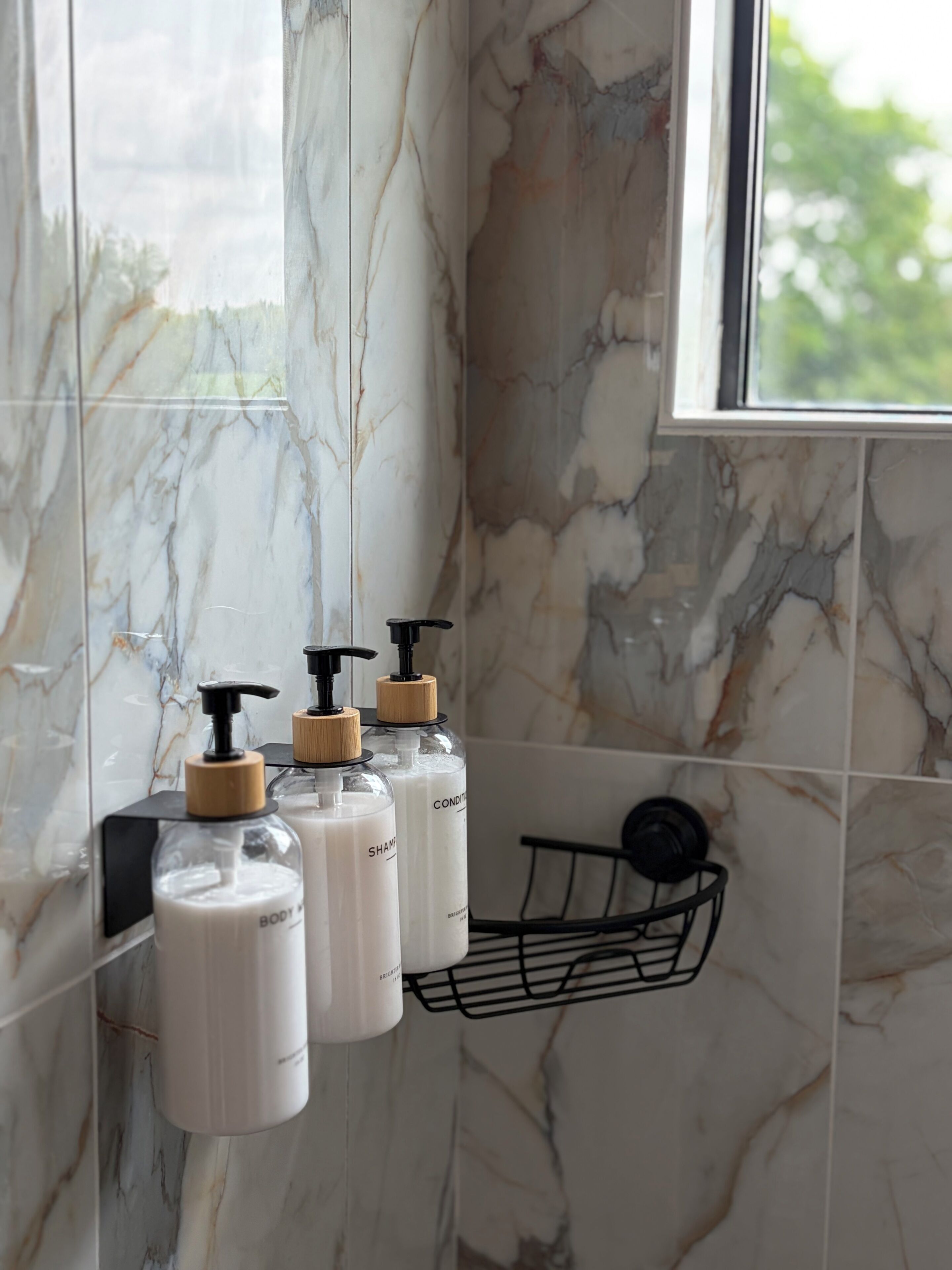 A bathroom with marble walls, a window, and three bottles of shampoo and conditioner.