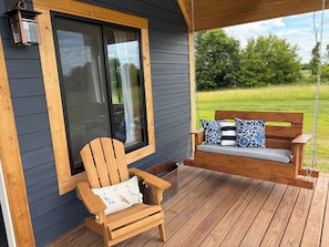 Une terrasse en bois avec une balançoire et une chaise, une fenêtre donnant sur un champ verdoyant et une lanterne fixée au mur.