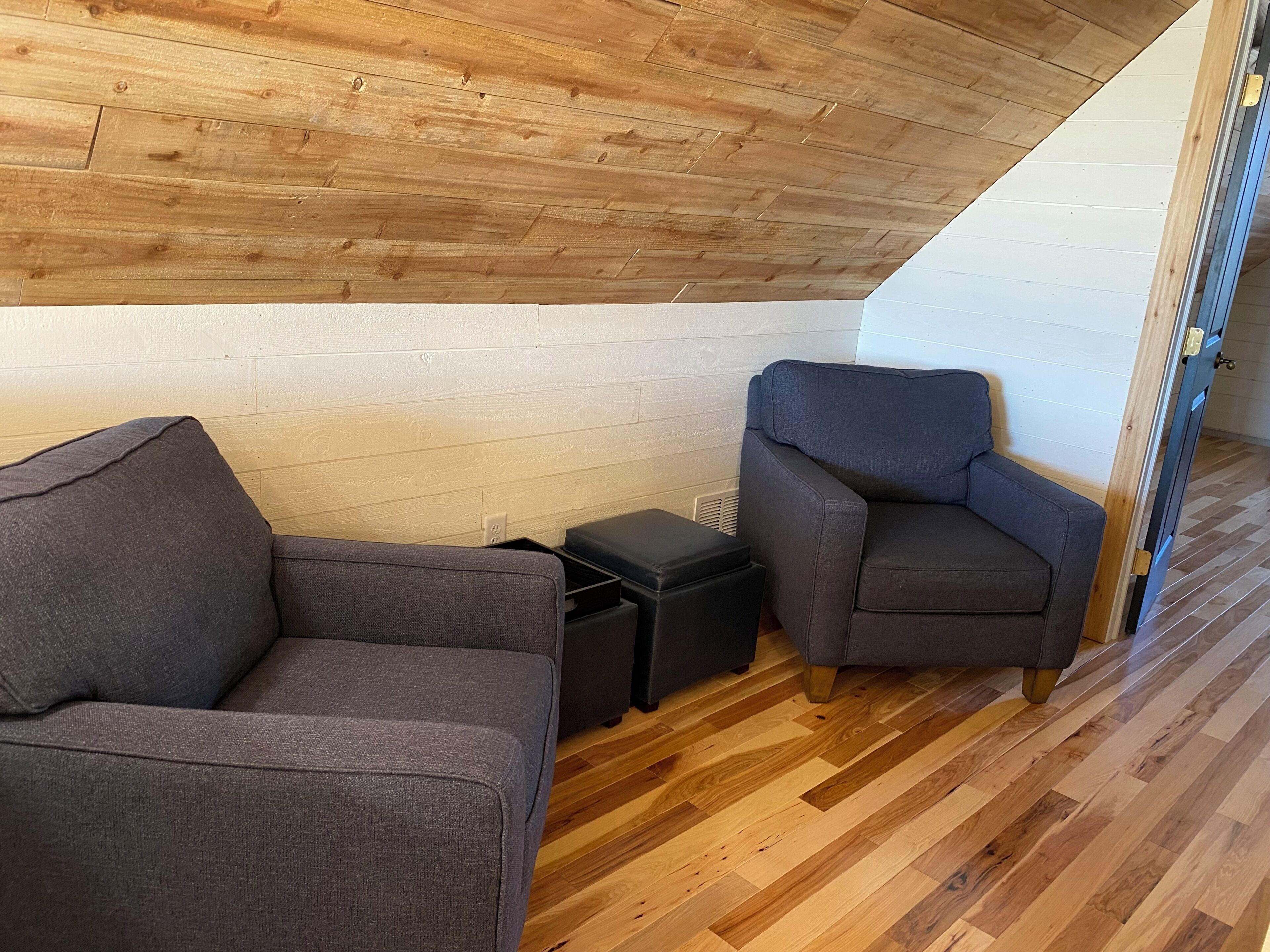 Una habitación con techo de madera, un sofá gris, un sillón gris y un pequeño mueble negro.