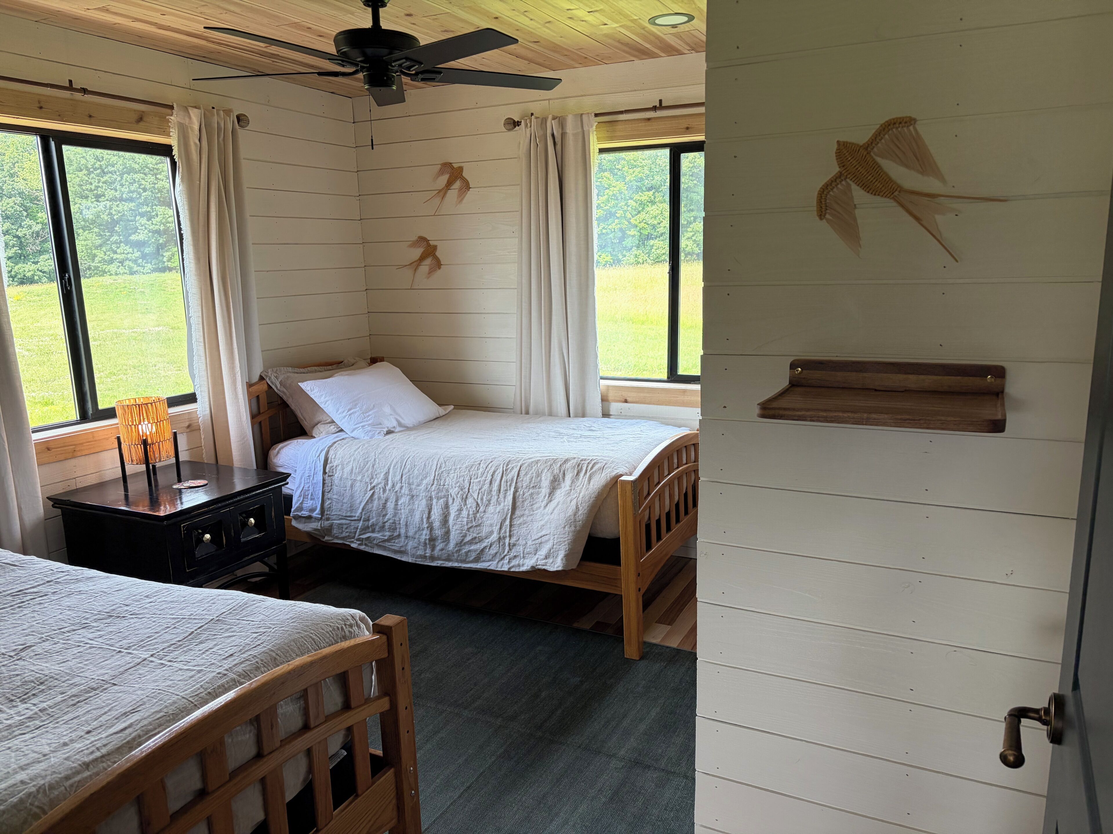 A room with two beds, a ceiling fan, a window with curtains, and a wall with a wooden shelf.