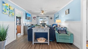 Ein Zimmer mit blauer Wand, einem Deckenventilator, einem Fernseher, einem Schreibtisch mit Korb und einem türkisfarbenen Sessel mit Strandkissen.