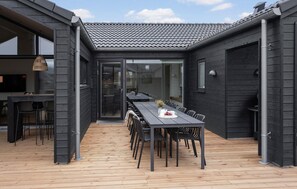 A modern outdoor dining area with a long table and chairs, a wooden deck, and a building with dark siding and a tiled roof. - Nice home in Otterup with sauna (Otterup)