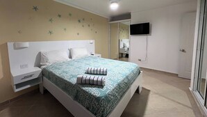 A bedroom with a bed, nightstands, a ceiling fan, a TV mounted on the wall, and a bathroom visible through an open door. - Garden Condos 48 - Friendly 1BR Apt, Sosua Center! (Sosúa)