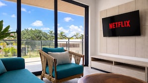 Ein modernes Wohnzimmer mit einem Korbstuhl, einem blaugrünen Sofa und einem Holztisch. Ein Flachbildfernseher an der Wand zeigt 'NETFLIX'.