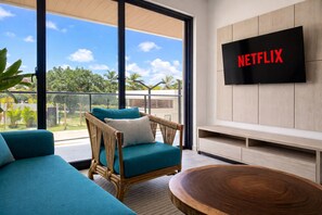Ein modernes Wohnzimmer mit einem Korbstuhl, einem blaugrünen Sofa und einem Holztisch. Ein Flachbildfernseher an der Wand zeigt 'NETFLIX'.