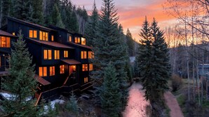 A multi-story building with large windows and a wooden exterior, surrounded by dense forest and a path leading to a cabin. - Riverstone Retreat by Avantstay Shared Hot Tub + Mountain Views Walk to Gondola + San Miguel River Trail (Telluride)