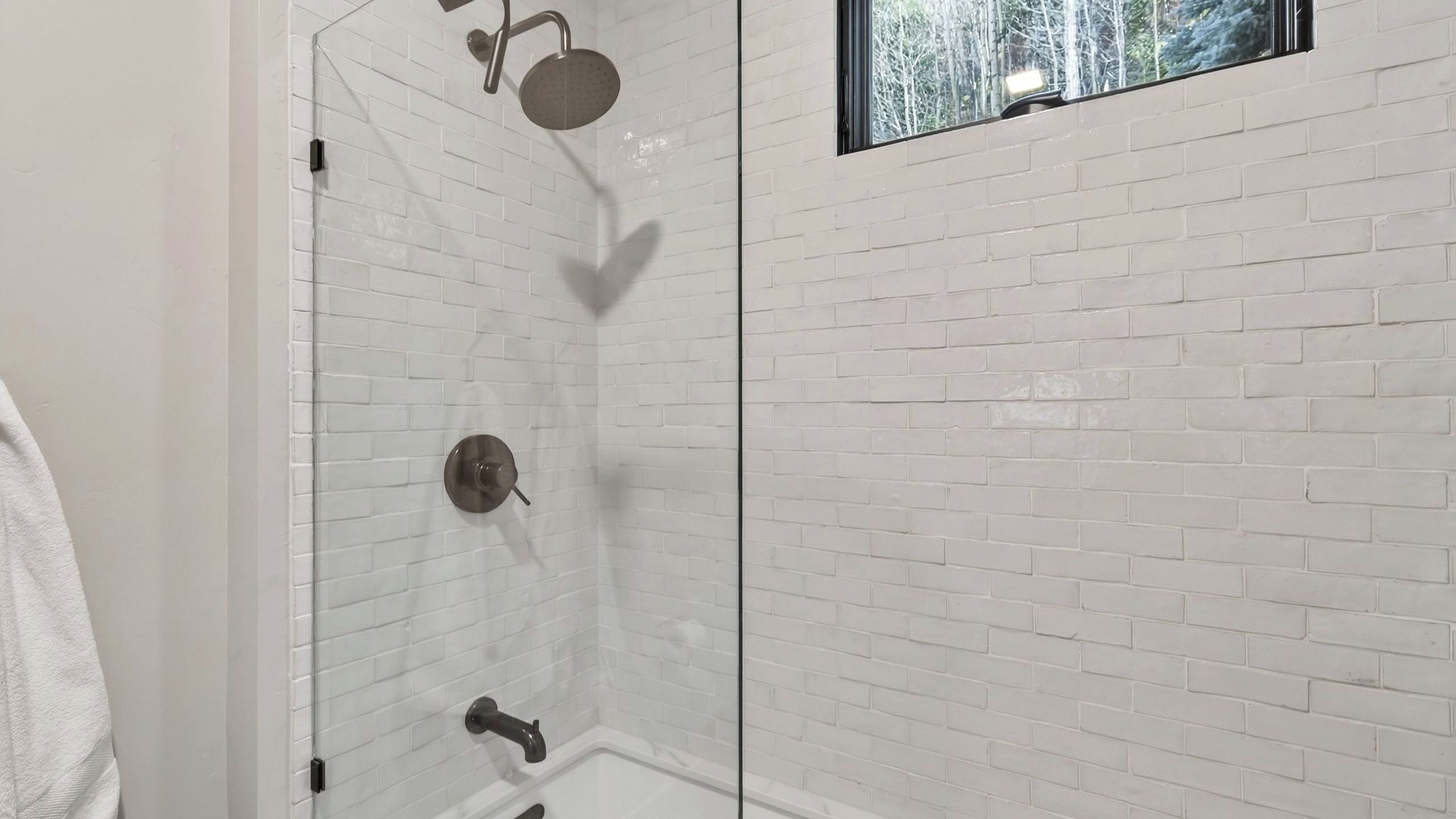 A shower with a glass door, white brick walls, and a window with a view of trees.