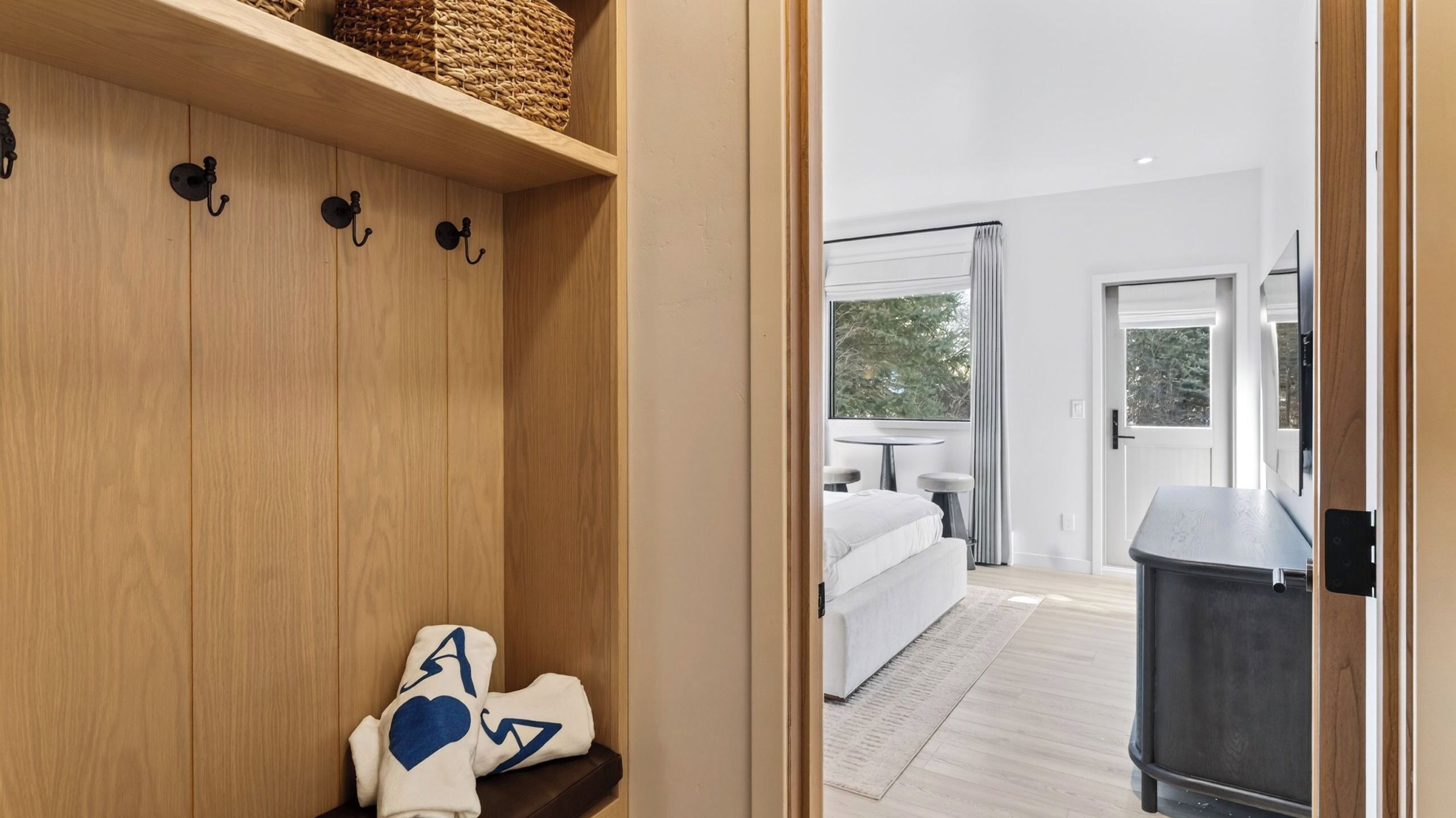 A wooden wardrobe with hooks, a bench with a towel, and a view into a bedroom with a bed and a window.