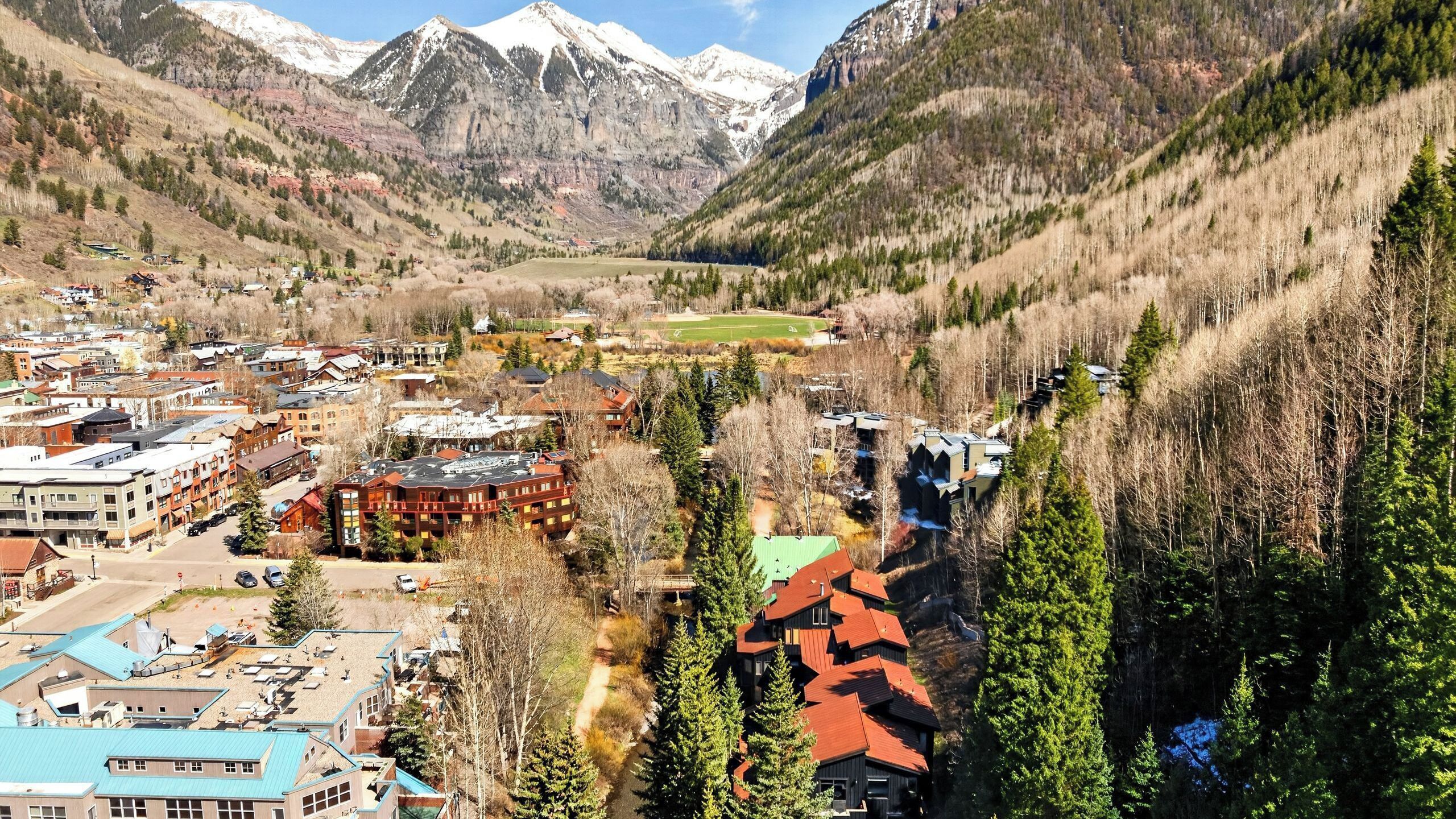 A mountainous town with buildings, a green field, and snow-capped peaks.