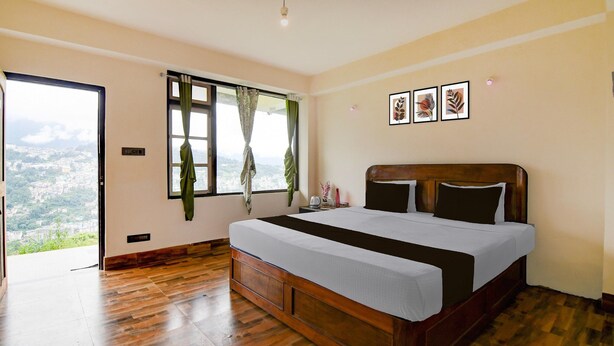 A bedroom with a wooden bed, a large window with curtains, and a city view. - Deluxe Hotel O Ranka Gangtok (Gangtok)