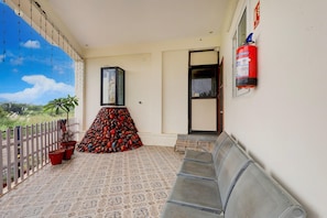 Un balcone con pavimento decorato, una fila di sedie in metallo, un estintore, un quadro incorniciato e un recinto bianco.