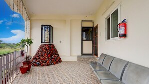 A balcony with a patterned floor, a row of potted plants, a statue, a fire extinguisher, and a seating area.