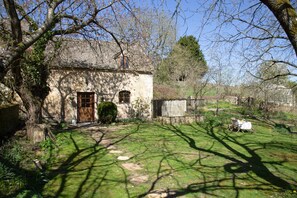 Ein Steinhaus mit einer Holztür, ein kleiner Garten mit Tisch und Stühlen und ein Baum, der Schatten auf das Gras wirft.