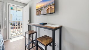 A room with a TV mounted on the wall, a wooden table with two stools, and a view of the beach through a door. - Paws & Palms-Pet Friendly-Pool-Free Parking (North Myrtle Beach)